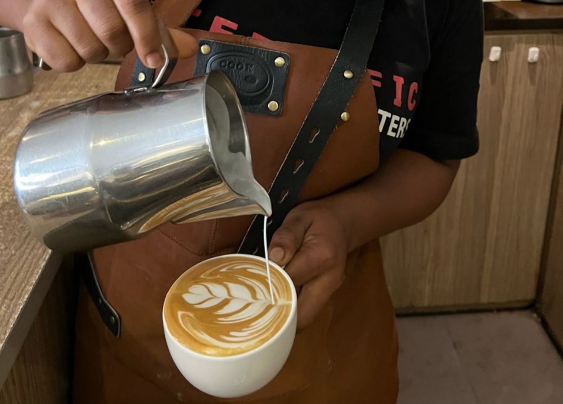 Barista gießt aufgeschäumte Milch aus Edelstahlkännchen in Kaffeetasse und kreiert Latte-Art-Muster